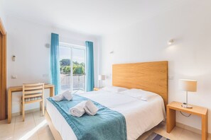 A hotel room with a bed, a desk, a chair, a nightstand, and a window with curtains. - Lumina Villas (Lagoa)