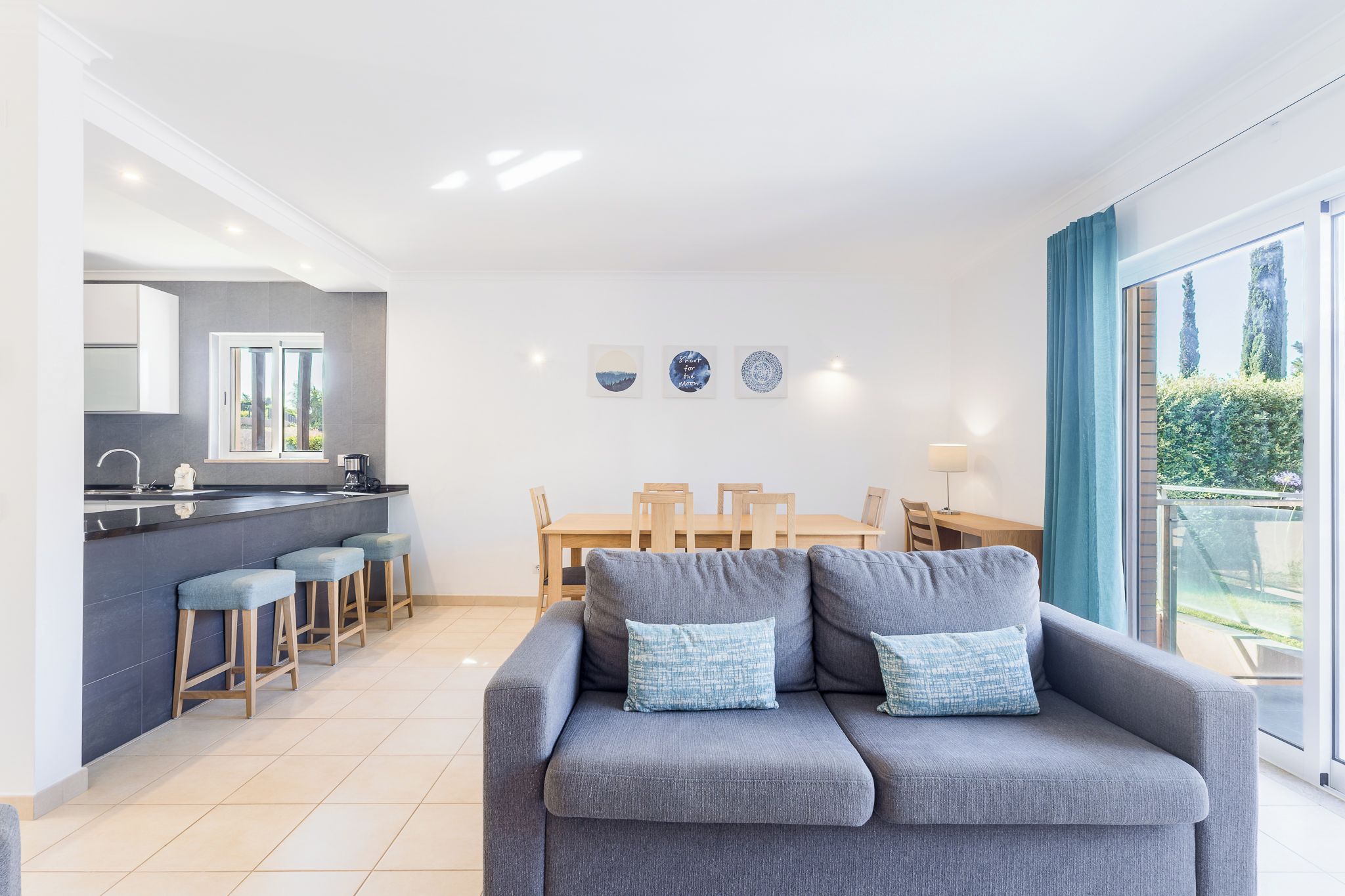 A modern living room with a grey sofa, a kitchen area with a bar, and a dining set.