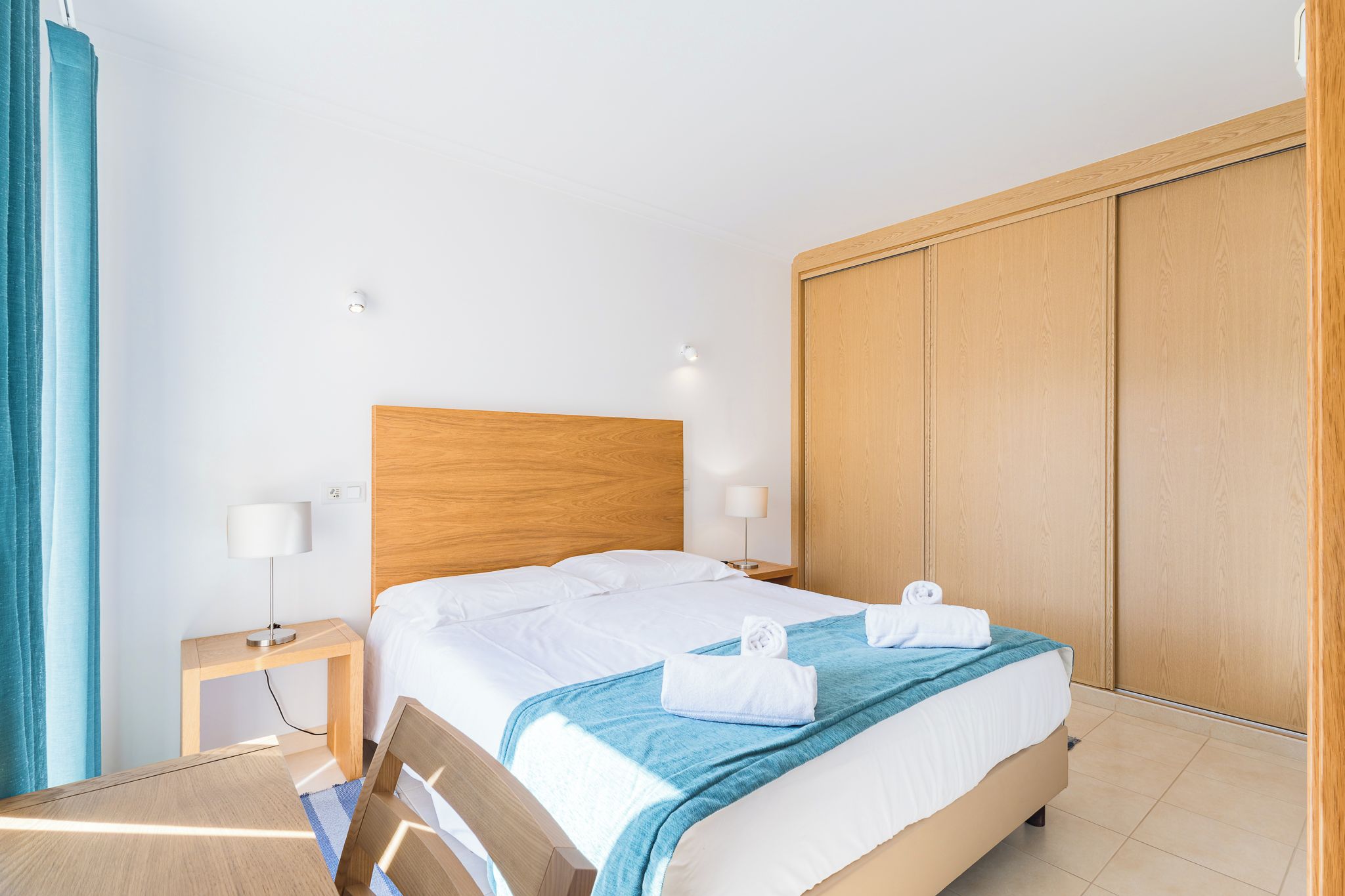 A hotel room with a wooden headboard, a bed with white linens, a blue throw blanket, and a wooden wardrobe.
