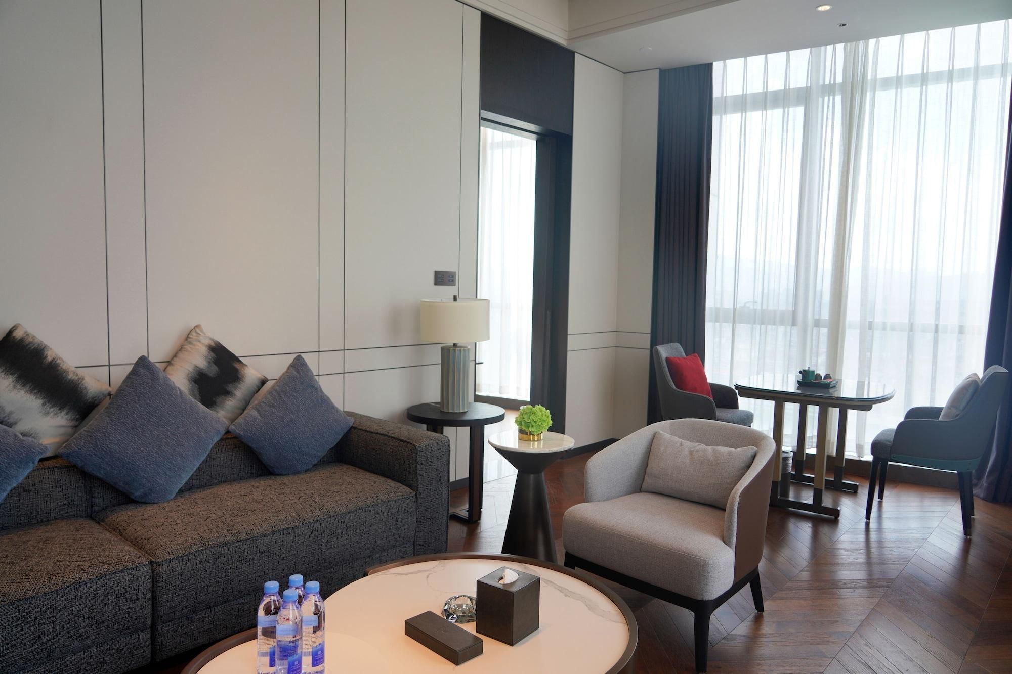 A modern hotel room with a sofa, armchair, small round table, and a dining area with chairs.