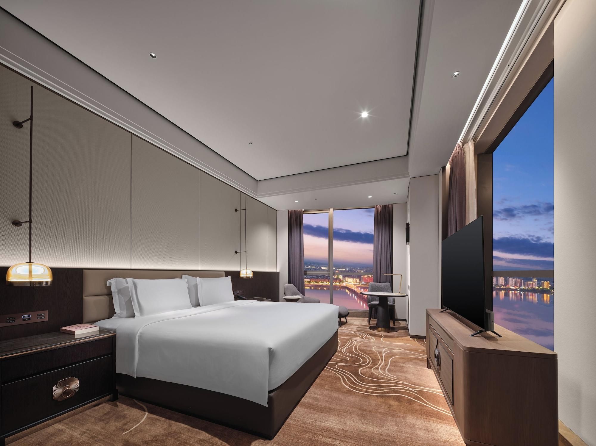 A modern hotel room with a large bed, a TV, a desk, and a view of a cityscape.