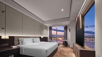 A modern hotel room with a large bed, a TV, a desk, and a view of a cityscape.