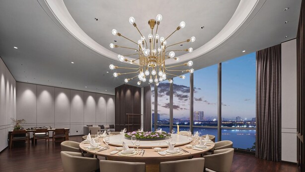 A modern dining room with a round table set for six, a chandelier, and a view of a cityscape and water. - Wanda Realm Yudu Ganzhou (Guangzhou)