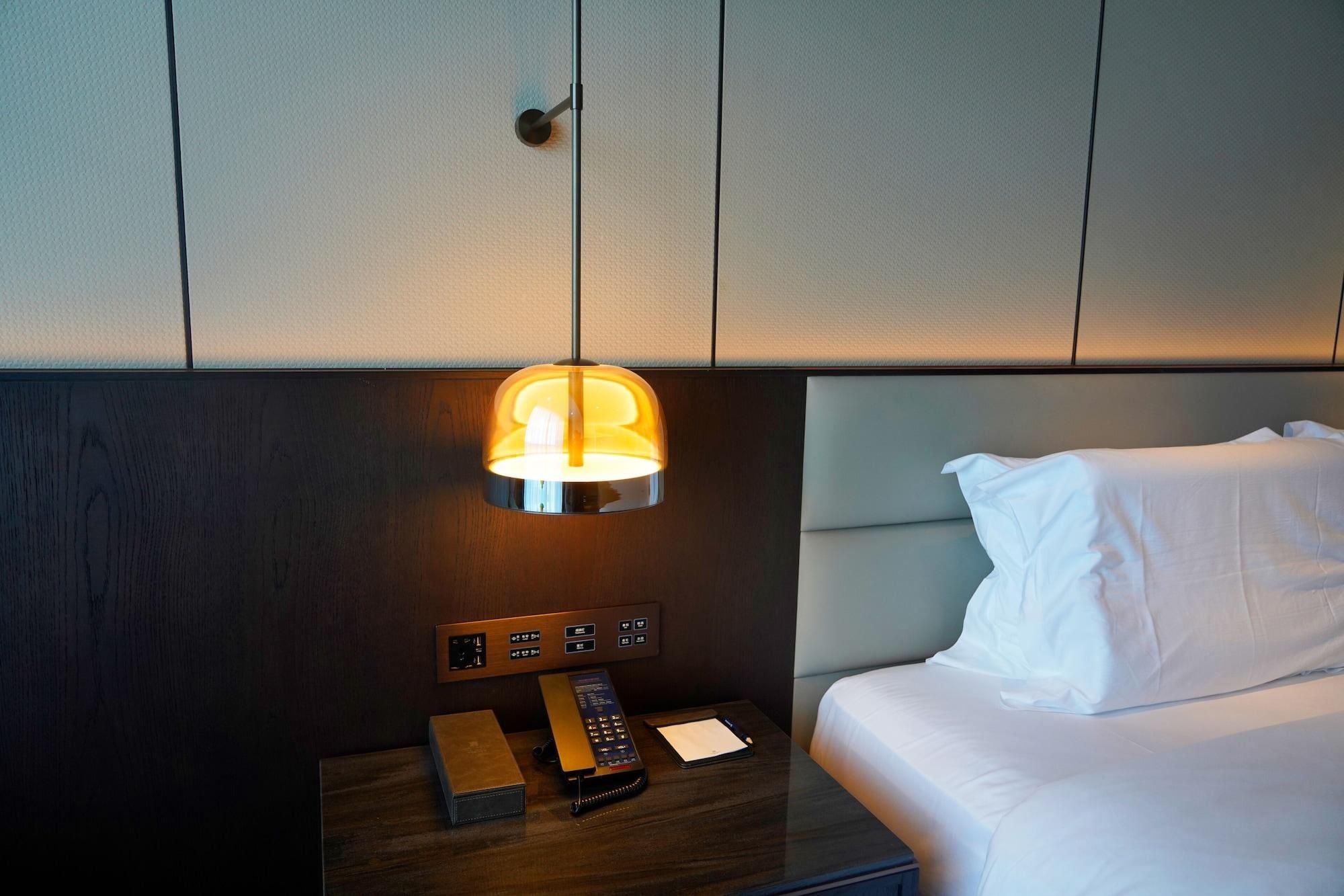 Et hotelværelse med en seng, et natbord med lampe, en fjernbetjening og en lille æske.