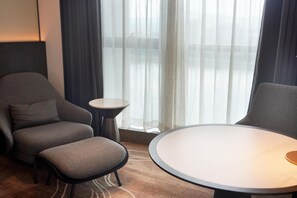 Una habitación de hotel con un sillón gris, una pequeña mesa redonda y un amplio ventanal con cortinas translúcidas.