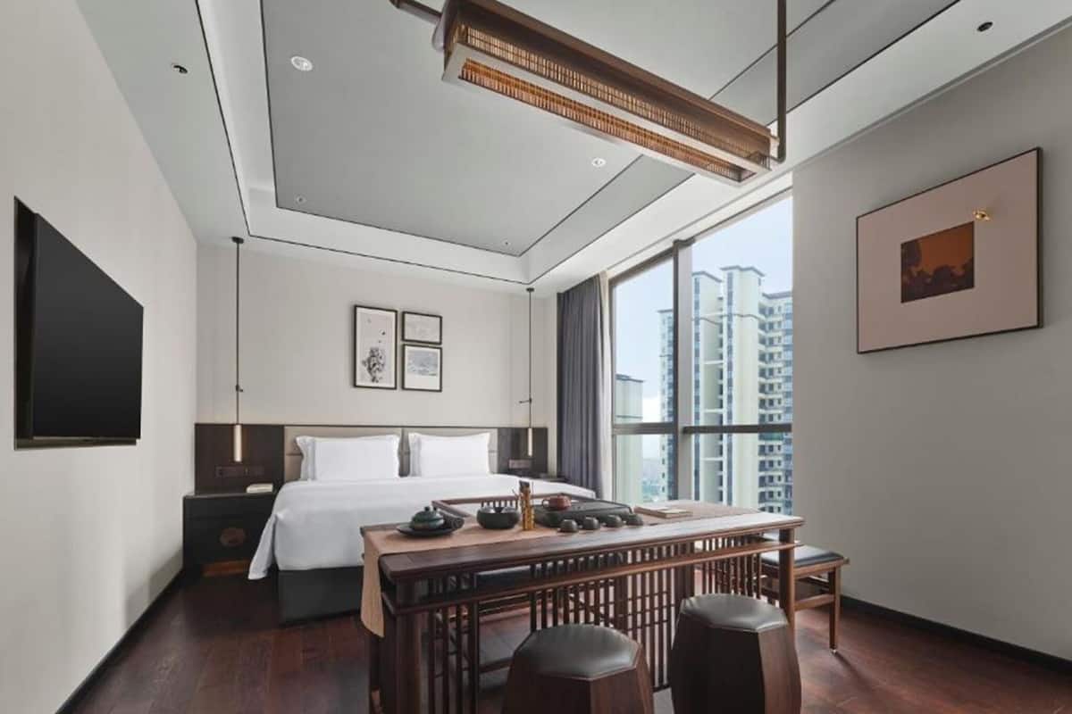 A modern hotel room with a large bed, a desk with stools, a flat-screen TV, and a large window offering a city view.