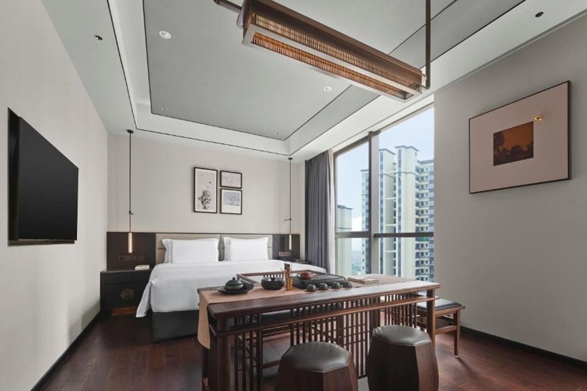 Una habitación de hotel moderna con una cama grande, un escritorio con taburetes, un televisor de pantalla plana y un amplio ventanal con vistas a la ciudad.