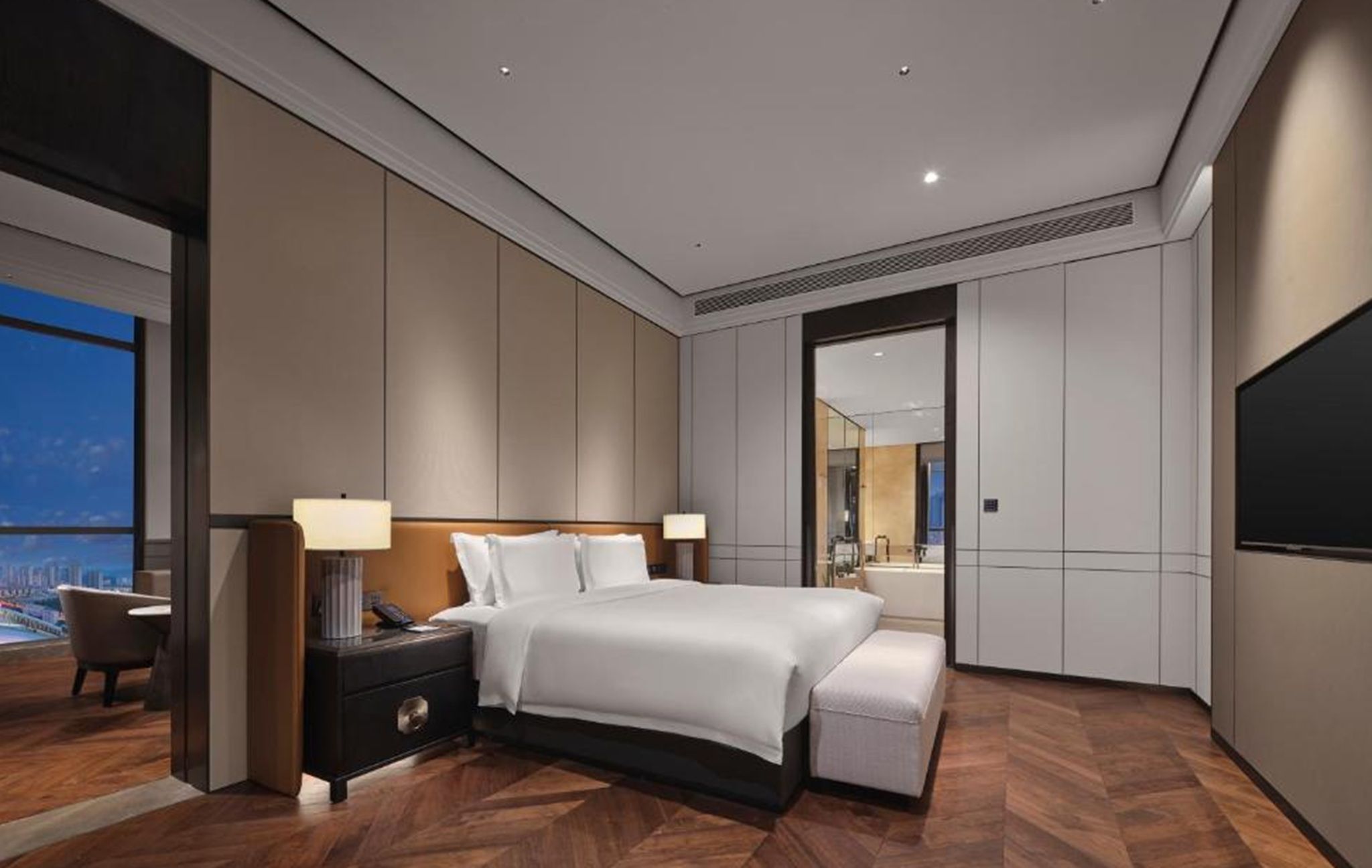 A modern hotel room with a large bed, bedside tables, a desk, a chair, and a large window offering a city view.