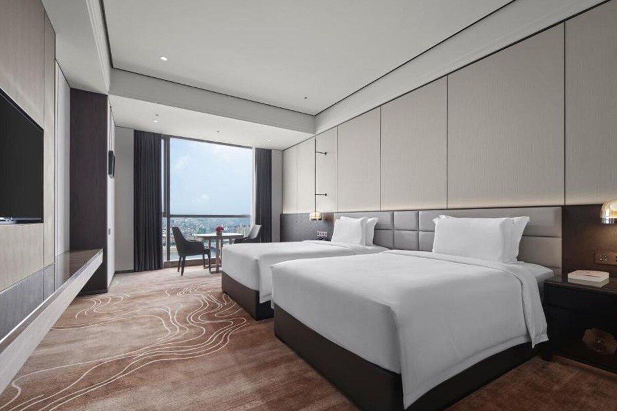 Habitación de hotel con dos camas, televisión, escritorio con silla y vista a la ciudad.