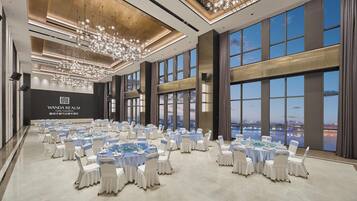 Une salle de banquet spacieuse, dotée de tables rondes, d’un lustre et de grandes fenêtres offrant une vue sur la ville.