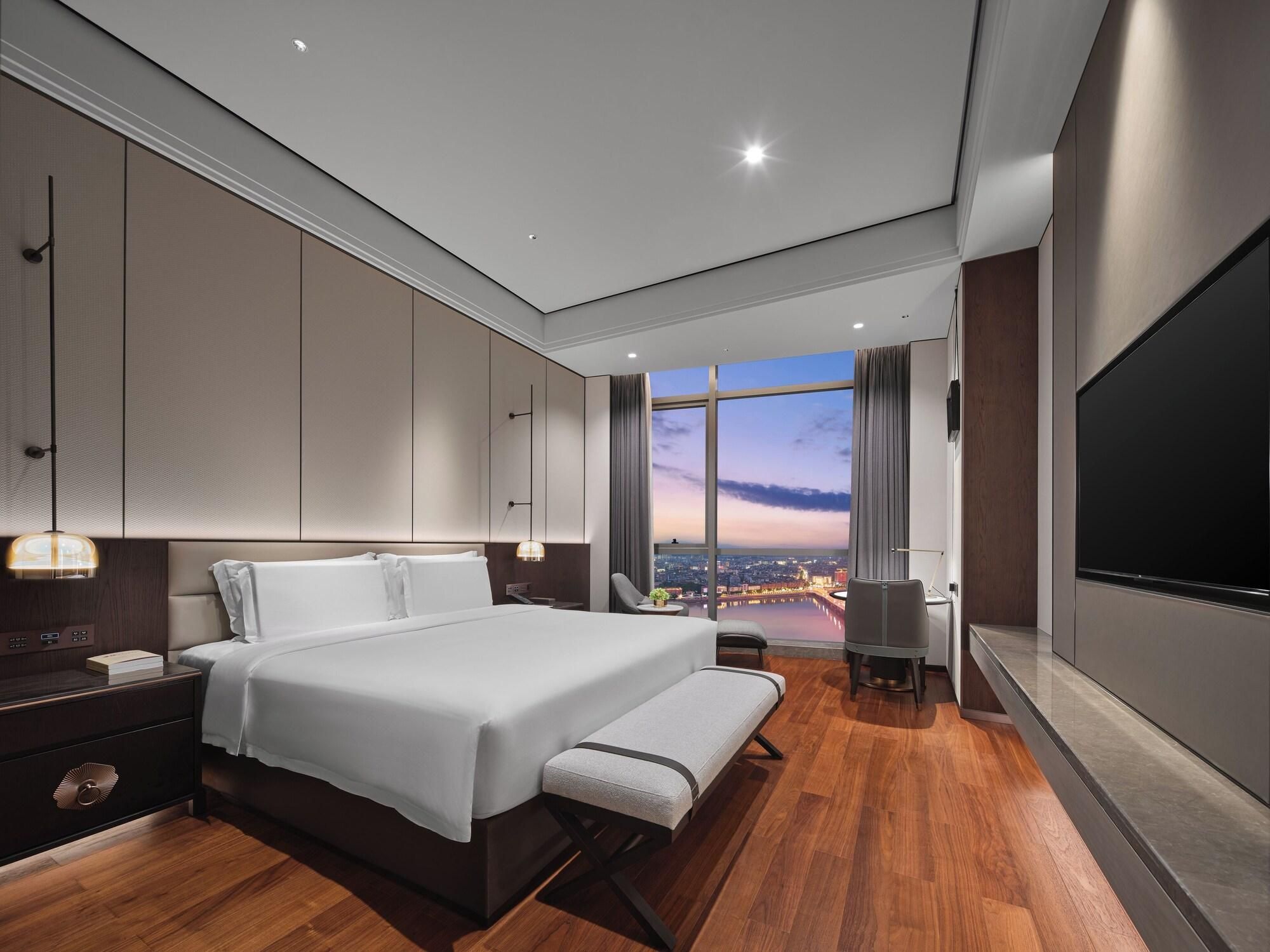 A modern hotel room with a large bed, a desk, a TV, and a view of the cityscape.
