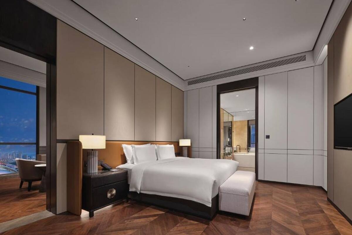 A modern hotel room with a large bed, bedside tables, a desk, a chair, and a large window offering a city view.