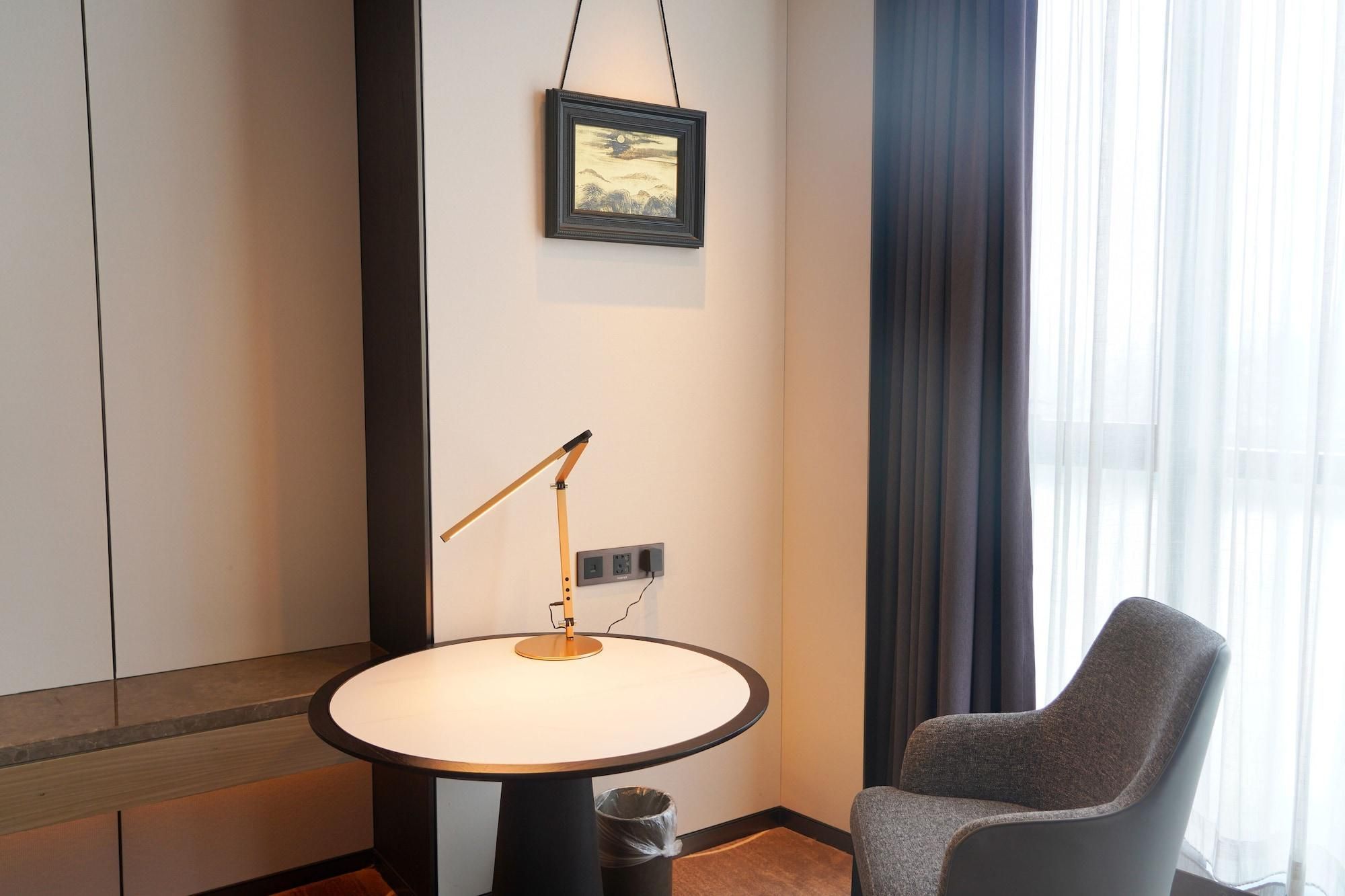 A hotel room with a round table, a single chair, a standing lamp, a power outlet, and a framed picture on the wall.