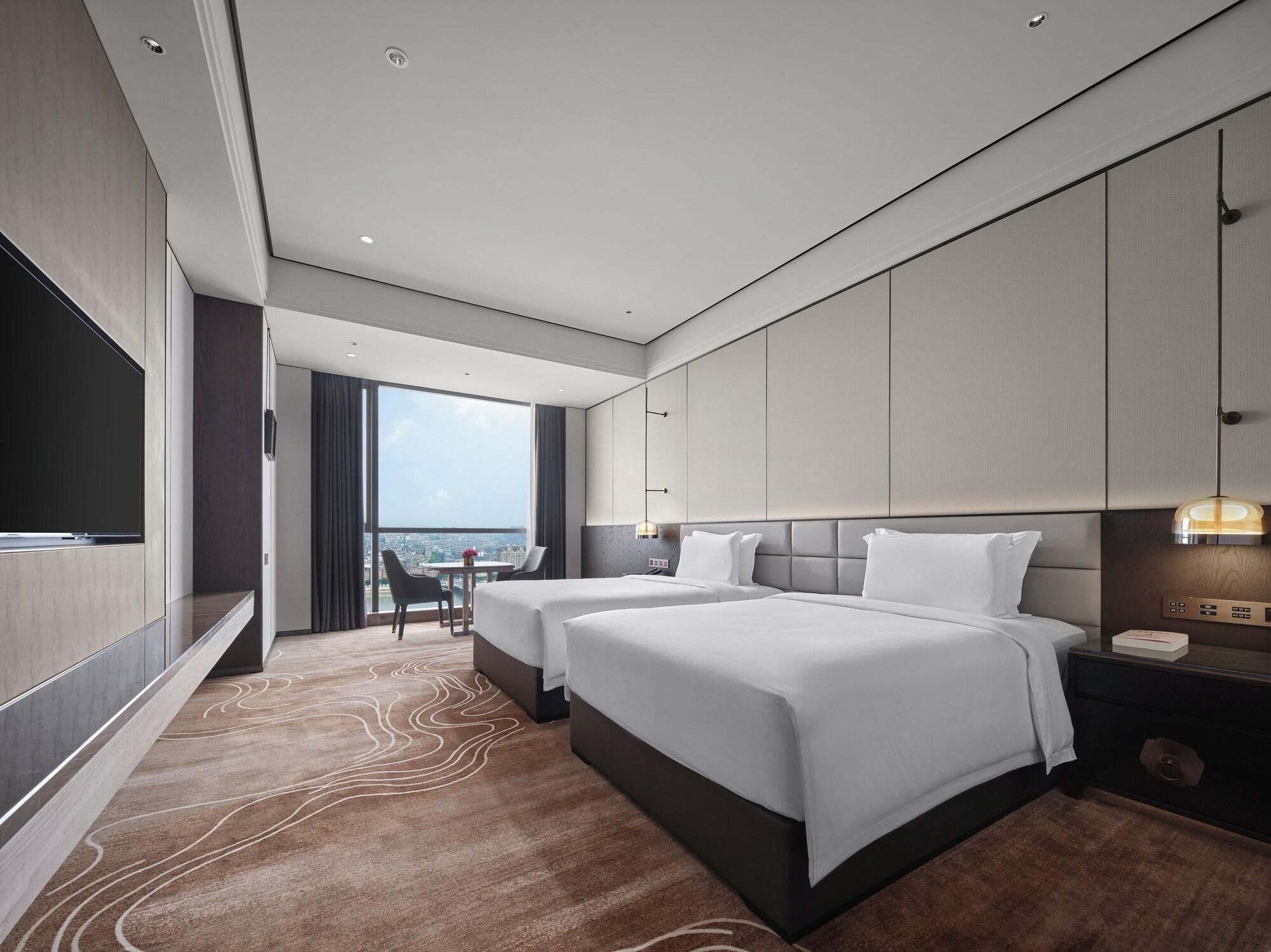 A modern hotel room with two beds, a TV, a desk, and a view of the city.