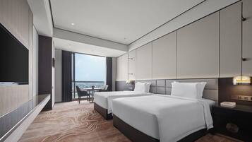 A modern hotel room with two beds, a TV, a desk, and a view of the city.