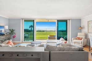 Ein modernes Wohnzimmer mit einem großen Schiebeglasfenster, das zu einer grünen Außenanlage führt, einer Couch, einem Couchtisch und einem Fernseher.