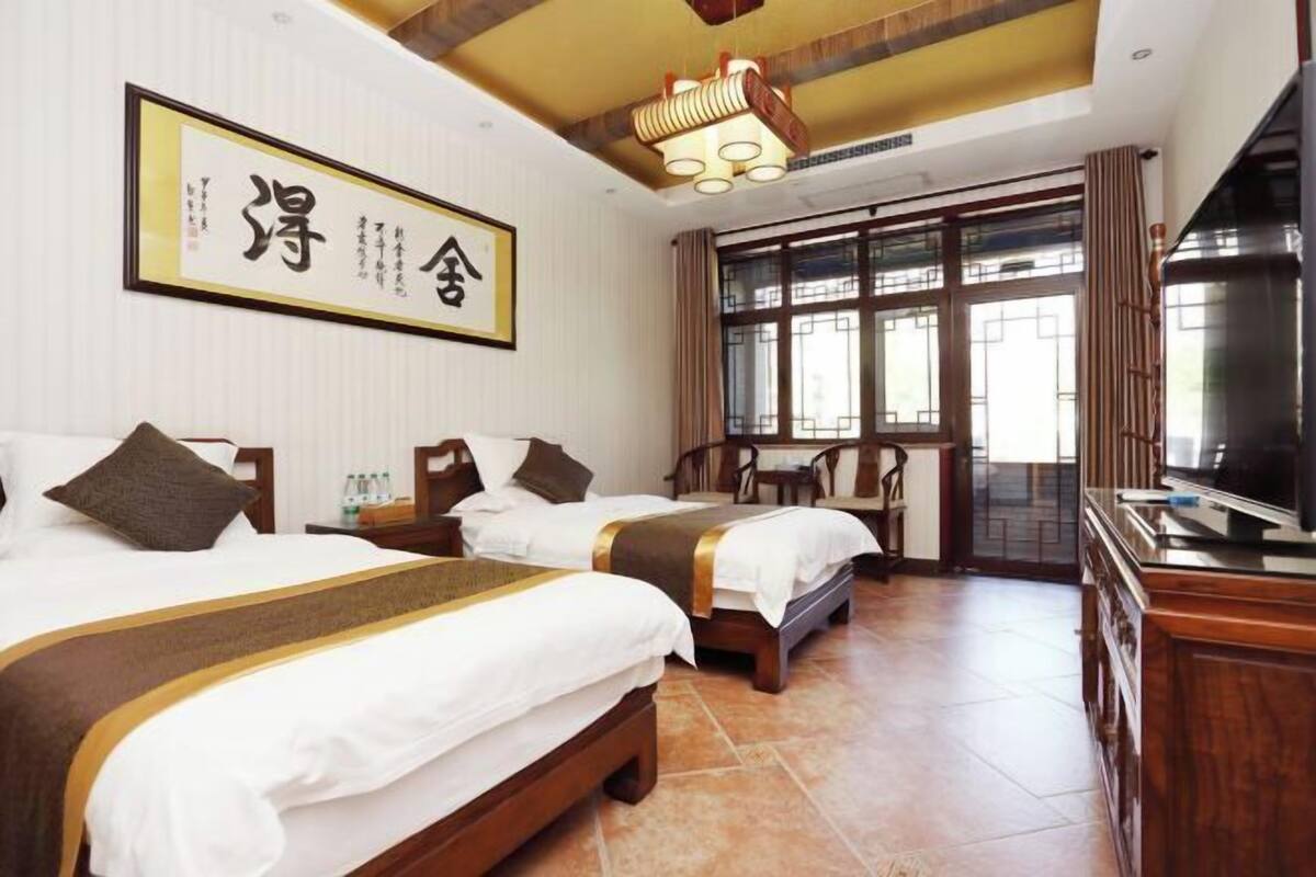 Quarto de hotel com três camas, televisão, armário de madeira e uma janela grande com cortinas.