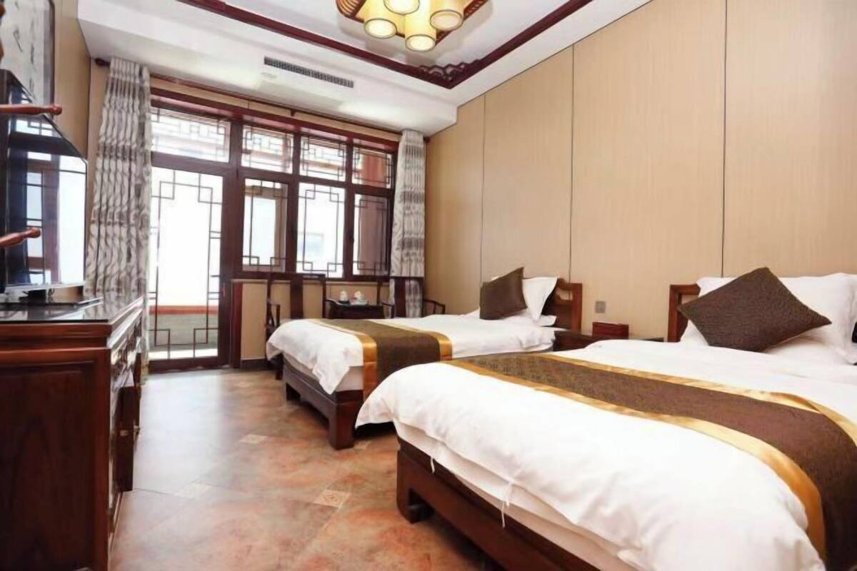 Quarto de hotel com duas camas, penteadeira de madeira, escrivaninha e uma janela grande com cortinas.