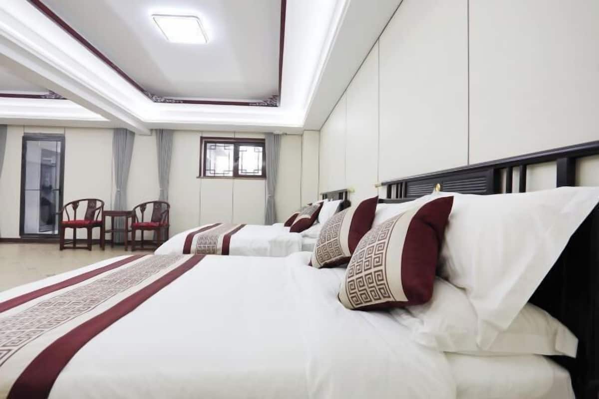 Quarto de hotel com três camas, área de estar com cadeiras e uma janela grande.