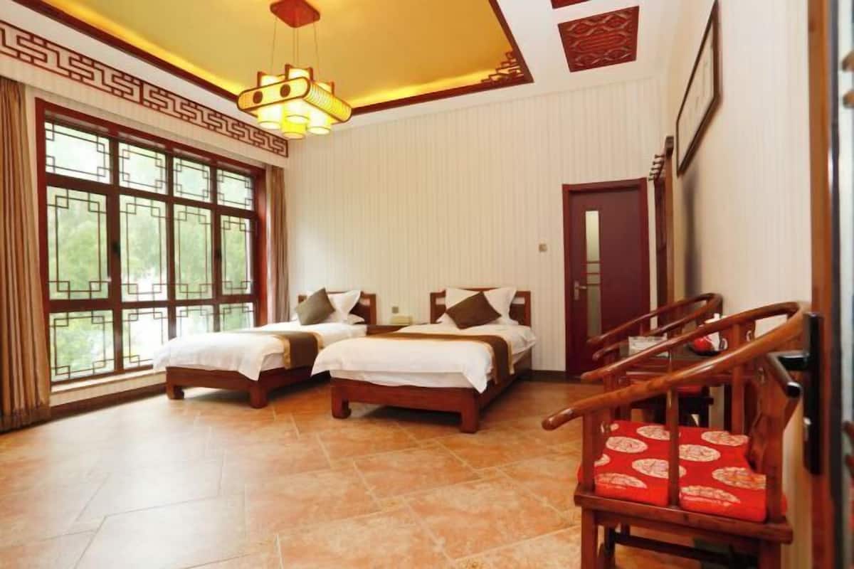 Quarto de hotel com duas camas, uma cadeira de madeira, uma janela grande e um design de teto tradicional.