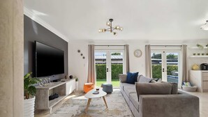 Ett modernt vardagsrum med en platt-TV, en grå soffa, ett soffbord och utsikt över en trädgård genom stora fönster.