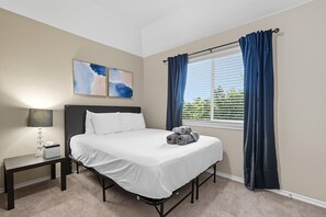 A neatly made bed with white linens, a bedside table with a lamp, and a window with blue curtains. - Spacious 5-bd/3-ba Forest View Getaway (Dallas)
