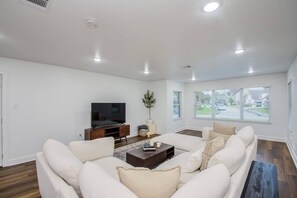 A modern living room with a large sofa, a coffee table, a flat-screen TV, and a window with a view of the street. - 3 Beds Houston Home, Close NRG Stadium & Rodeo (Houston)