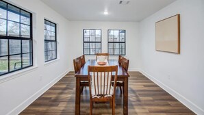 Un espace repas avec une table et des chaises en bois, de grandes fenêtres et un mur blanc.