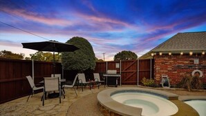 A backyard with a hot tub, patio furniture, and a brick building. - Modern, Spacious - Pool & Backyard Oasis & Cinema (Rowlett)