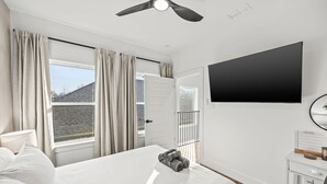 Ett hotellrum med en säng, en tv som är monterad på väggen, ett fönster med gardiner, en takfläkt och en dörr som leder ut till en balkong.