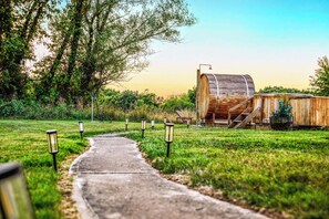 Un sendero conduce a una construcción de madera rústica con techo abovedado, rodeada de vegetación y un cielo despejado.