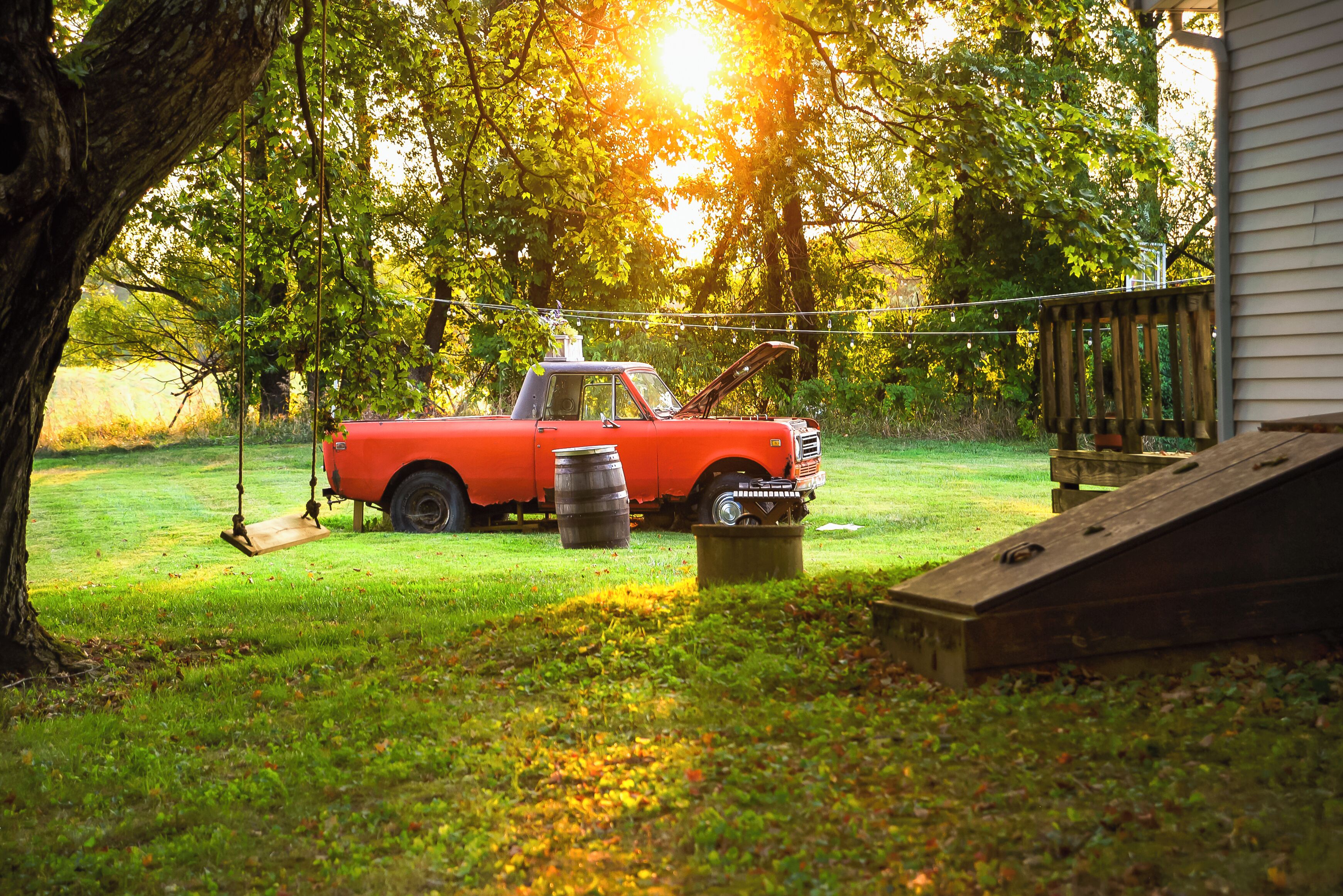 Un coche rojo antiguo estacionado en un patio con césped, una rampa de madera y un barril metálico.