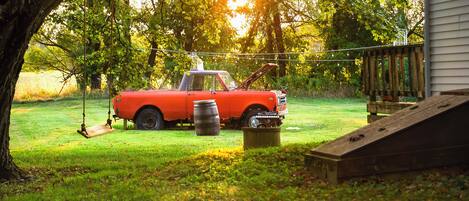 Un coche rojo antiguo estacionado en un patio con césped, una rampa de madera y un barril metálico.