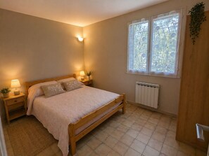 Ein Schlafzimmer mit einem hölzernen Bett, Nachttischlampen, einem Heizkörper und einem Fenster mit Blick auf Grünflächen.