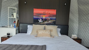 A bedroom with a large bed, a headboard with a painting, a bedside table with a lamp, and a nightstand with a tissue box. - Riverfront Dome Retreat | Private Bath + Views (Calgary)