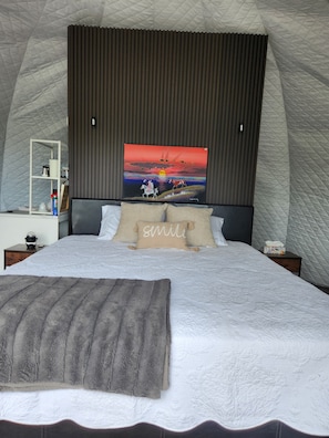 A bedroom with a large bed, a headboard with a painting, a bedside table with a lamp, and a nightstand with a tissue box.