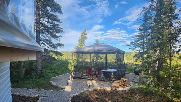 A covered outdoor area with a grill and seating, surrounded by trees and a gravel path. - Riverfront Dome Retreat | Private Bath + Views (Calgary)