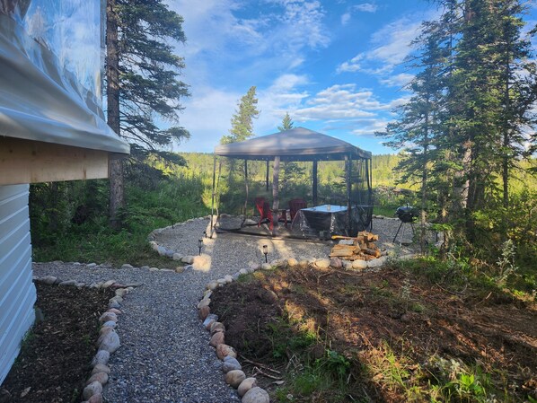 A covered outdoor area with a grill and seating, surrounded by trees and a gravel path.