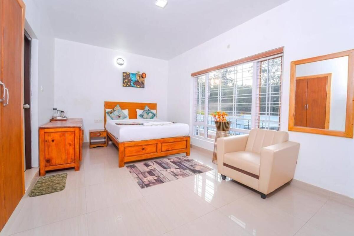 A bedroom with a bed, a wooden nightstand, a chair, a window with blinds, and a painting on the wall.