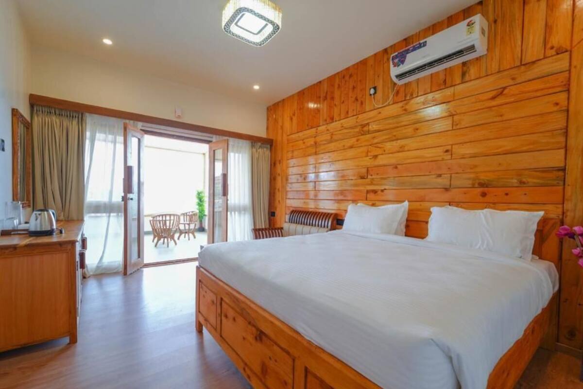 A hotel room with a wooden headboard, a large bed, a desk with a kettle, and a sliding glass door leading to a balcony.