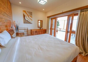 A bedroom with a large bed, wooden headboard, a desk, a chair, a mirror, and a sliding glass door leading to a balcony.