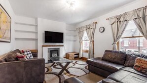 A living room with a fireplace, leather sofas, a flat-screen TV, and a large window with curtains. - Beautiful 3 Bedroom Flat in Fulham (London)