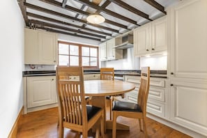 Une cuisine avec des chaises en bois, des armoires blanches, une table ronde et des poutres apparentes en bois au plafond.