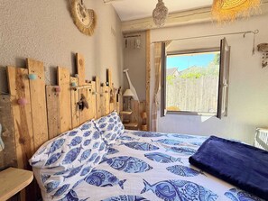 Ein Schlafzimmer mit hölzernem Kopfteil, einem Bett mit blauen Kissen und einer Tagesdecke mit Muster, einem Fenster mit Blick auf einen Zaun und einer Pendelleuchte.