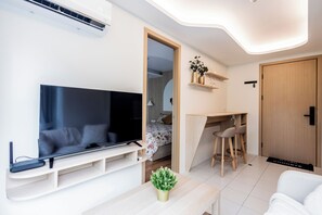 A modern living room with a flat-screen TV, a small table with a plant, a desk with a chair, and a bed with a floral bedspread. - The best vacation at Kamala Beach. Clean, friendly, and quiet 709 (Tambon Kamala)