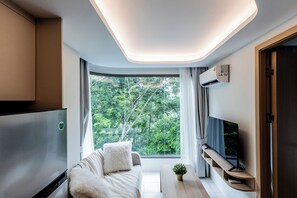 A modern living room with a sofa, a coffee table, a TV, and a large window with a view of trees. - The best vacation at Kamala Beach. Clean, friendly, and quiet 709 (Tambon Kamala)