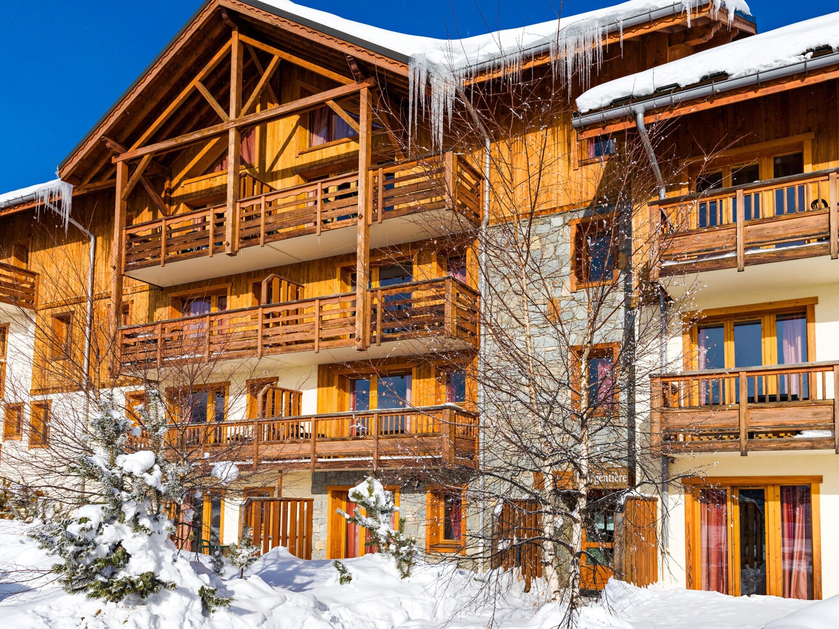 A multi-story wooden chalet with balconies, snow-covered surroundings, and a clear blue sky.