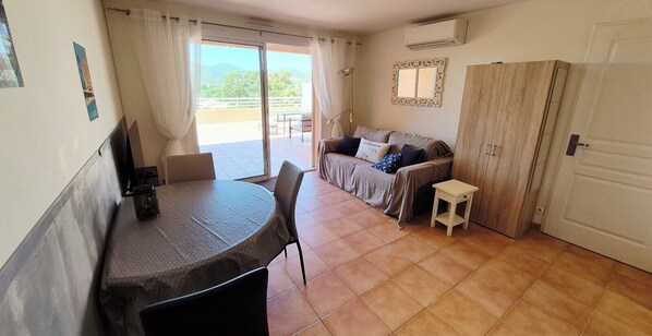 A room with a dining table, a sofa, a small stool, a wardrobe, and a sliding glass door leading to an outdoor area.