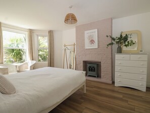 A bedroom with a bed, a dresser, a mirror, a fireplace, and a window with curtains. - East Cliff Gardens (Folkestone)