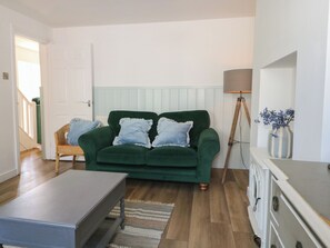 Un sofá verde con almohadones festoneados, piso de madera, una lámpara de pie y una silla de mimbre.